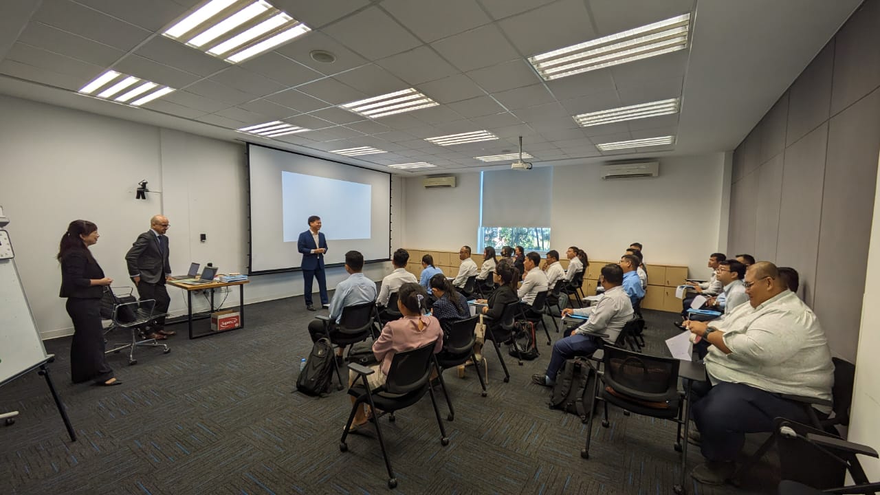 Indoor training: Man stands near screen, presenting to seated group. Two stand at side.